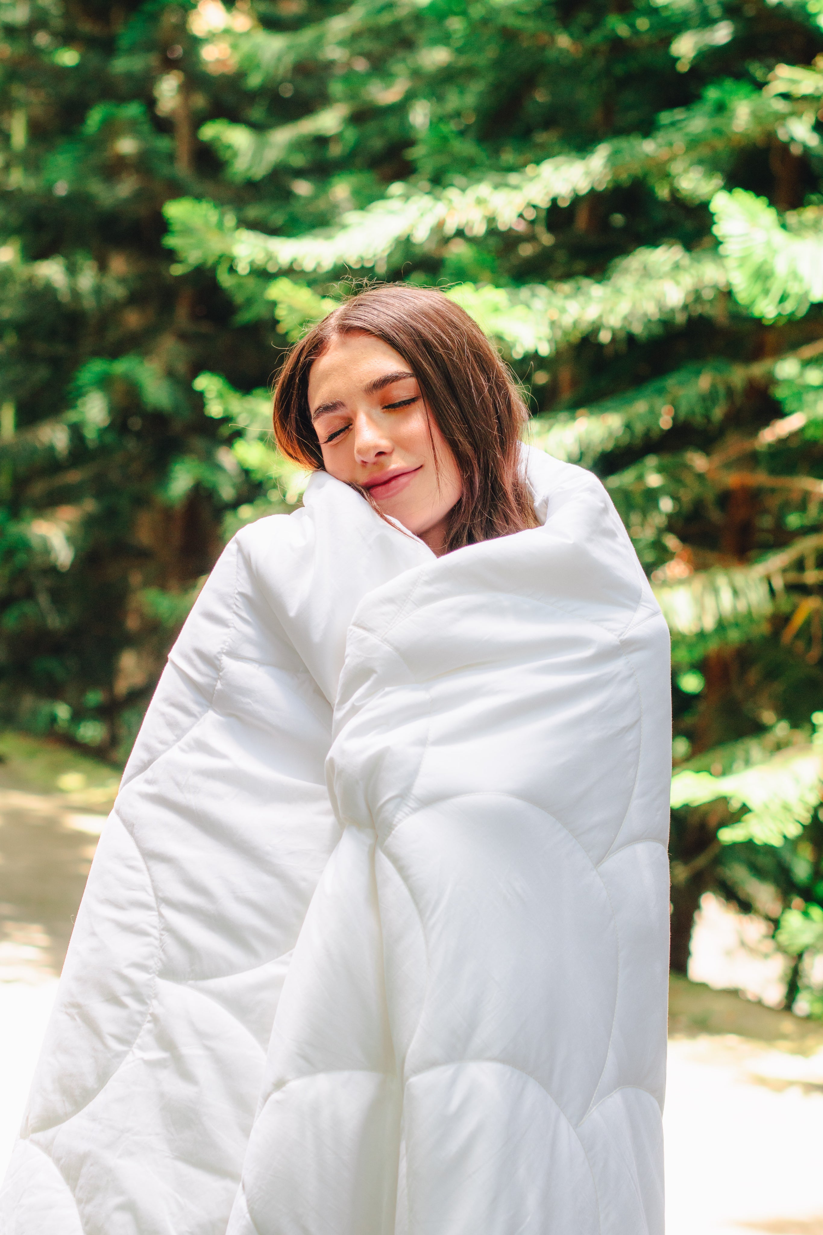 A woman wrapped in a white Larke duvet among evergreen trees